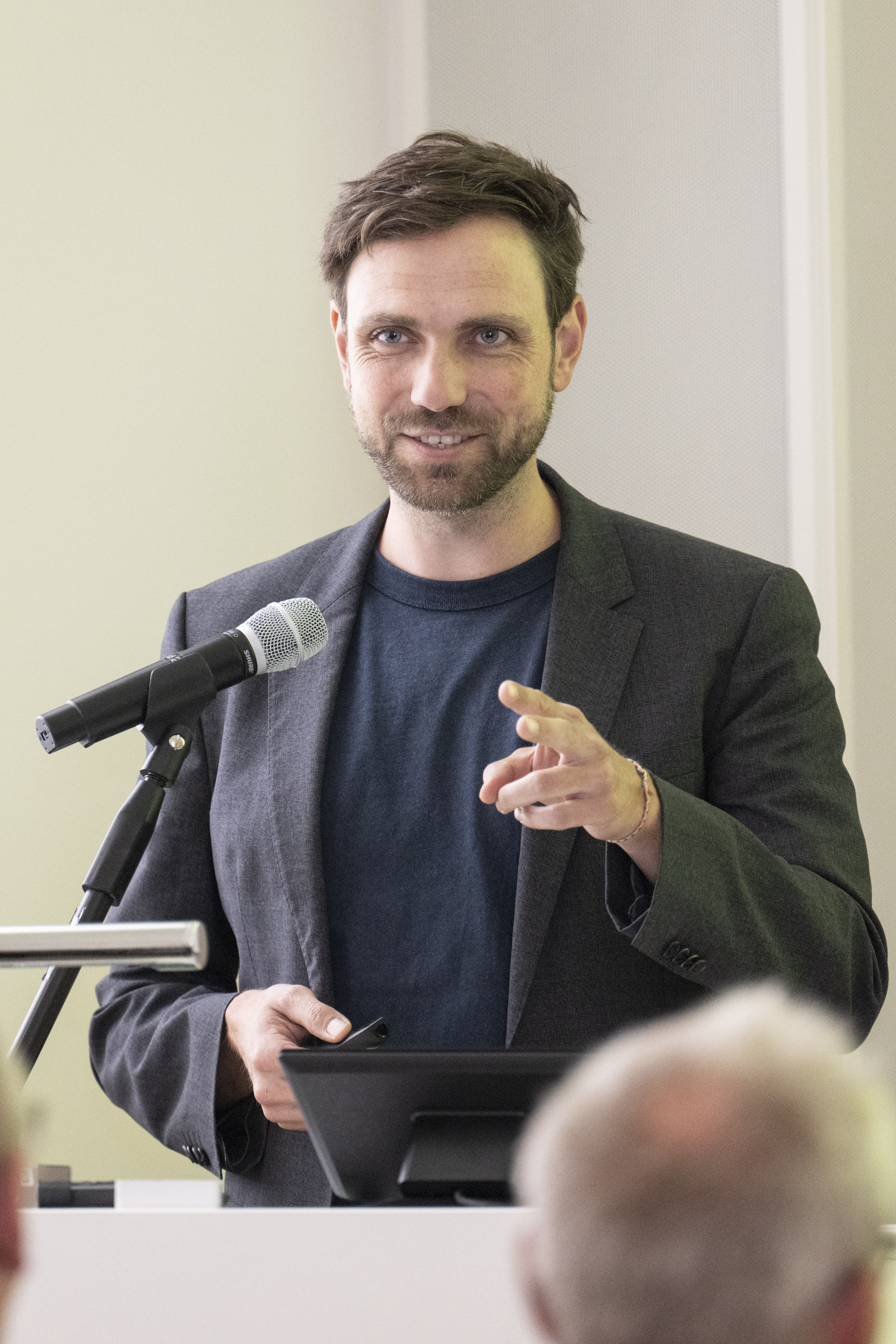 Man sieht Simon Wieland. Er hält einen Vortrag vor einer Menschengruppe